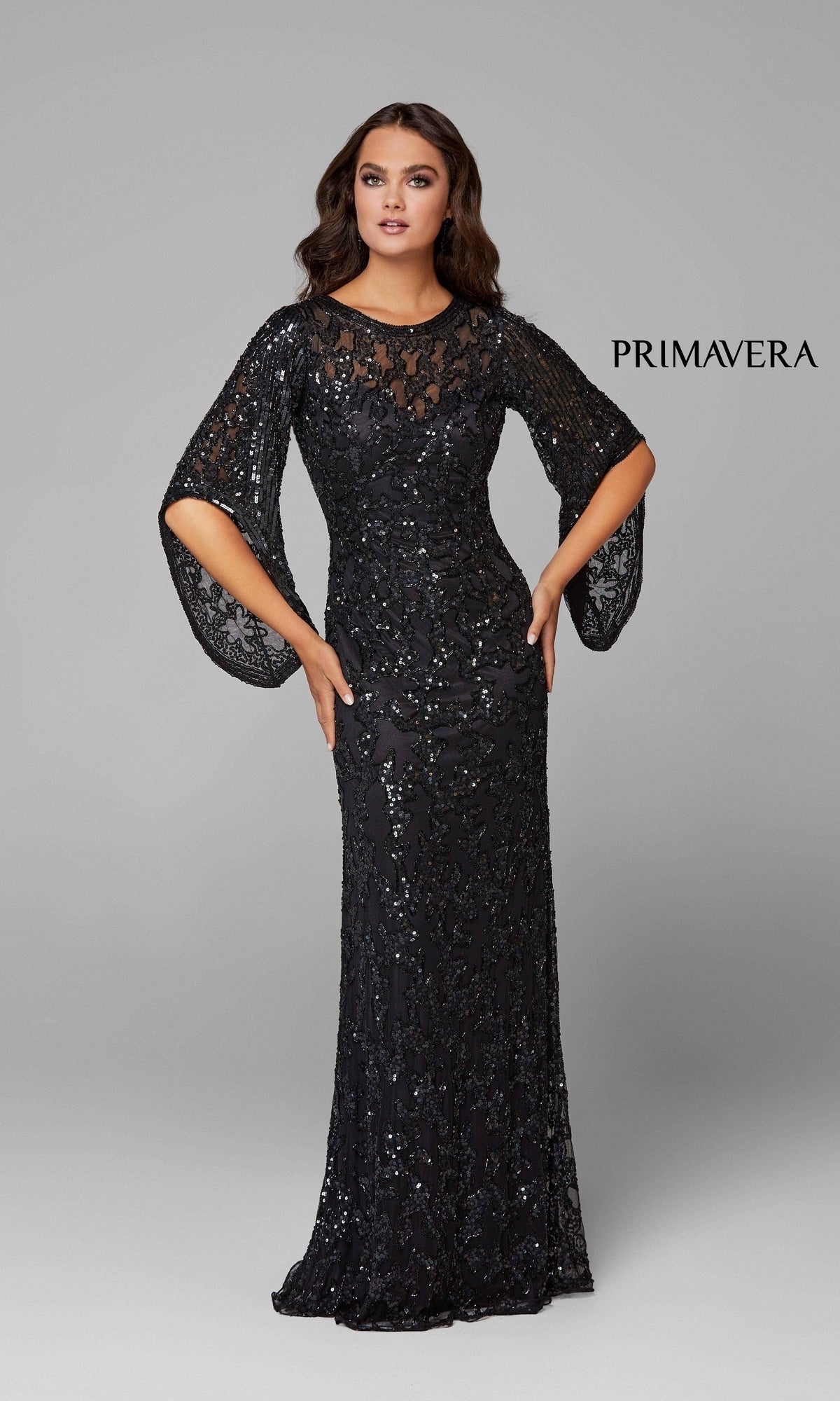 A woman models the Long Bell Sleeve Beaded Evening Gown 9713, a floor-length black dress with sheer, bell sleeves and intricate beading, against a plain gray background.